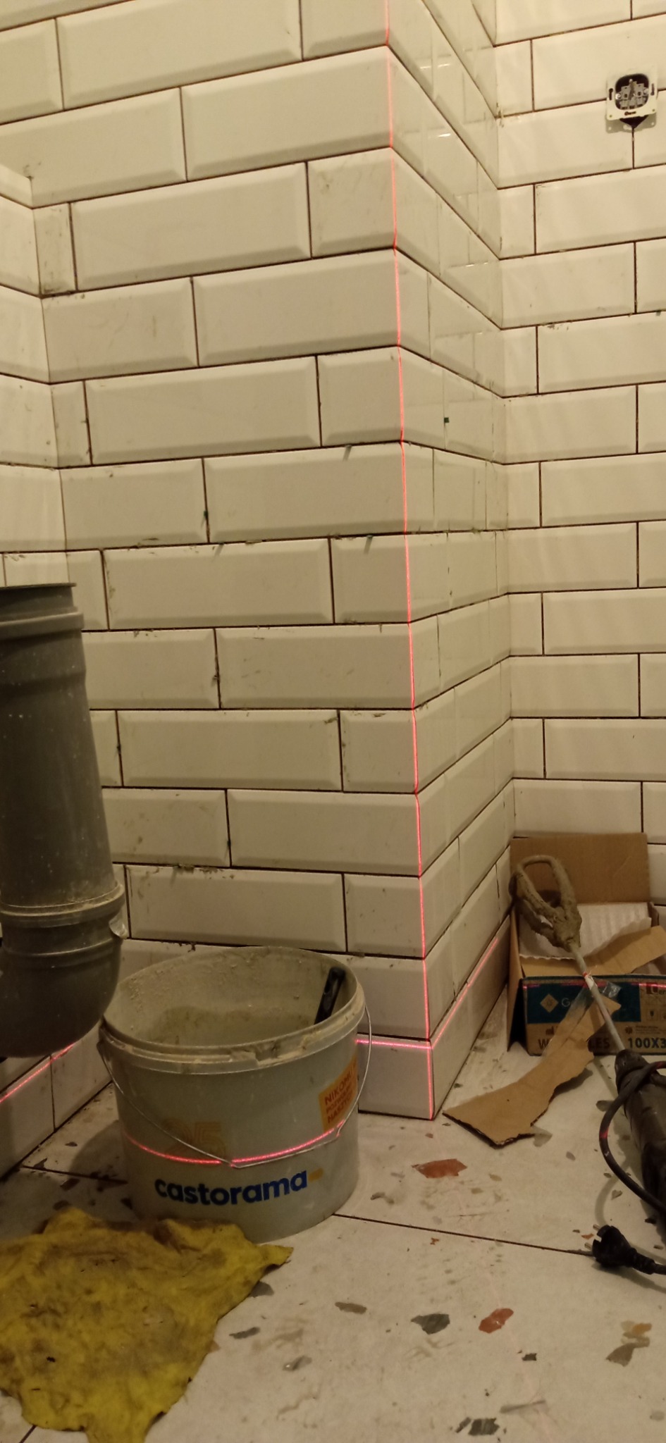 Ułożone białe, prostokątne płytki na ścianie, wyznaczanie poziomu czerwoną linią lasera, wiadro z zaprawą Castorama, widoczny odpływ kanalizacyjny i narzędzia na podłodze z lastryko.