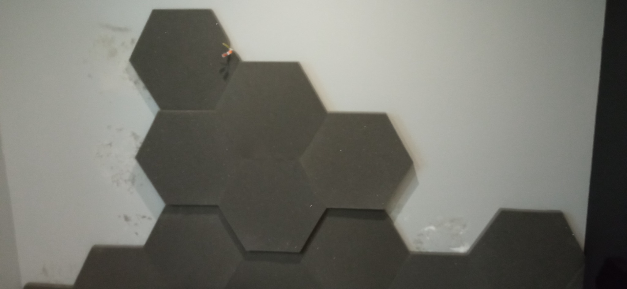 Ciemnoszare panele akustyczne w kształcie heksagonów umieszczone na jasnoszarej ścianie z widocznymi śladami kleju lub zabrudzeniami.