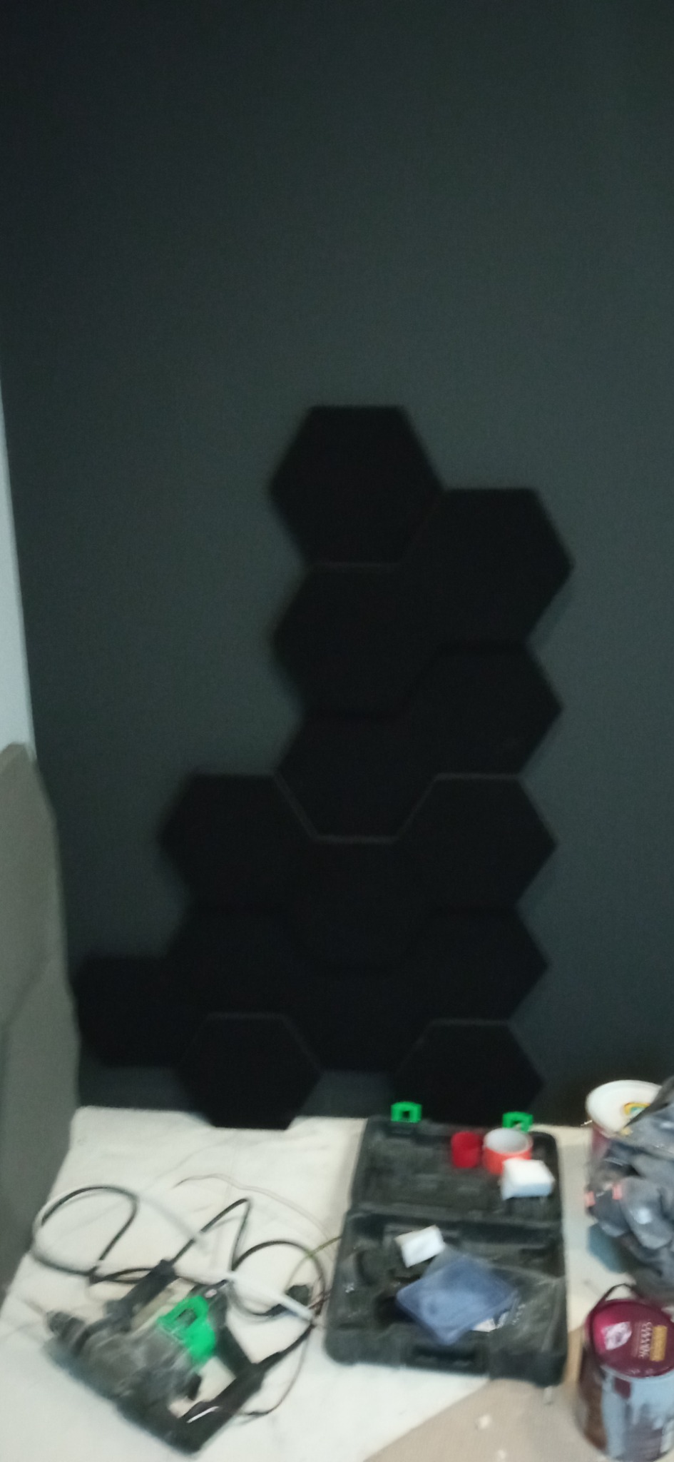 Montaż czarnych paneli akustycznych o heksagonalnym kształcie na szarej ścianie, widoczne narzędzia i puszki z farbą na podłodze.