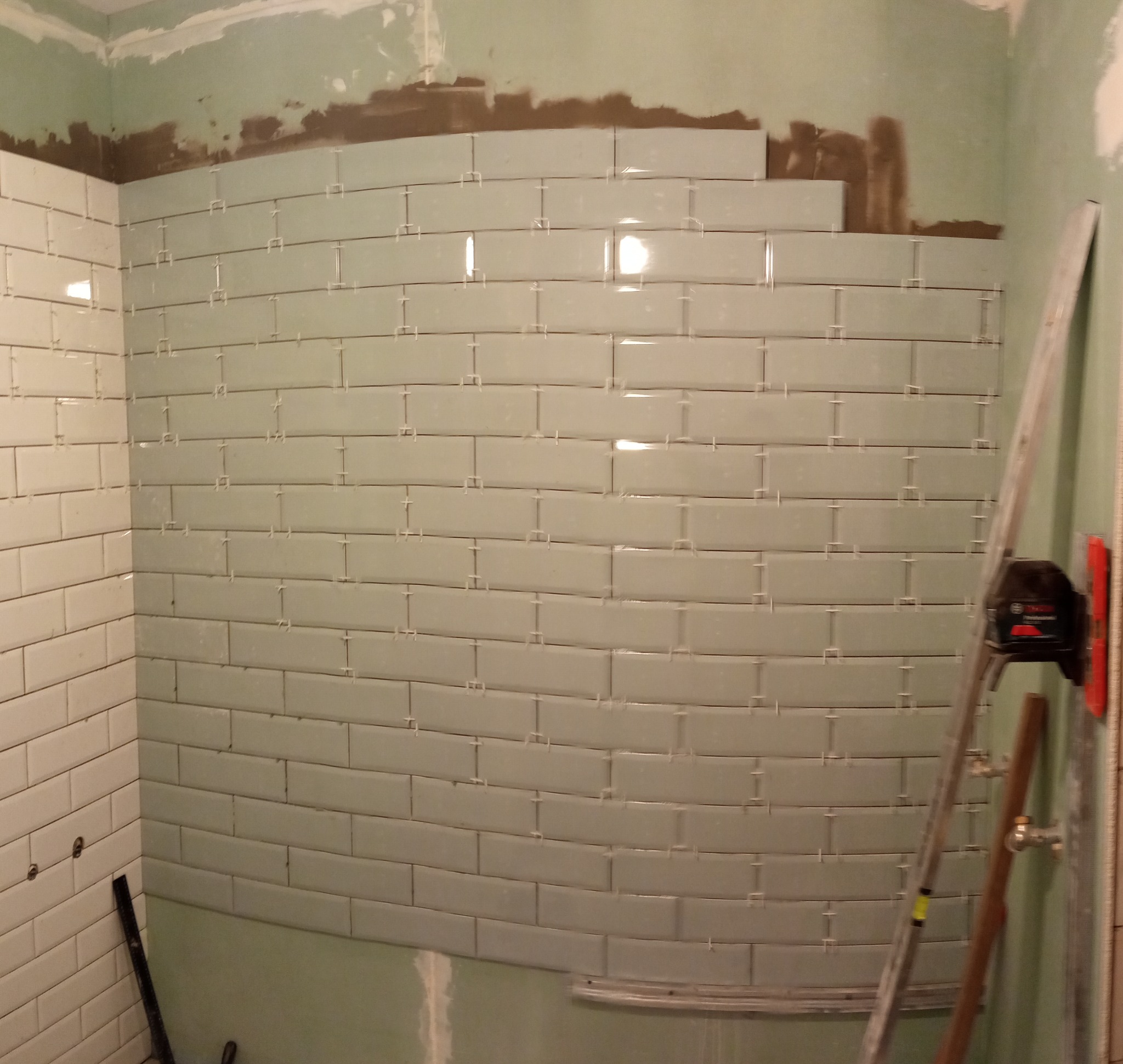 Układanie jasnych, prostokątnych płytek na ścianie w łazience, z użyciem krzyżyków dystansowych, widoczna drabina i poziomica.