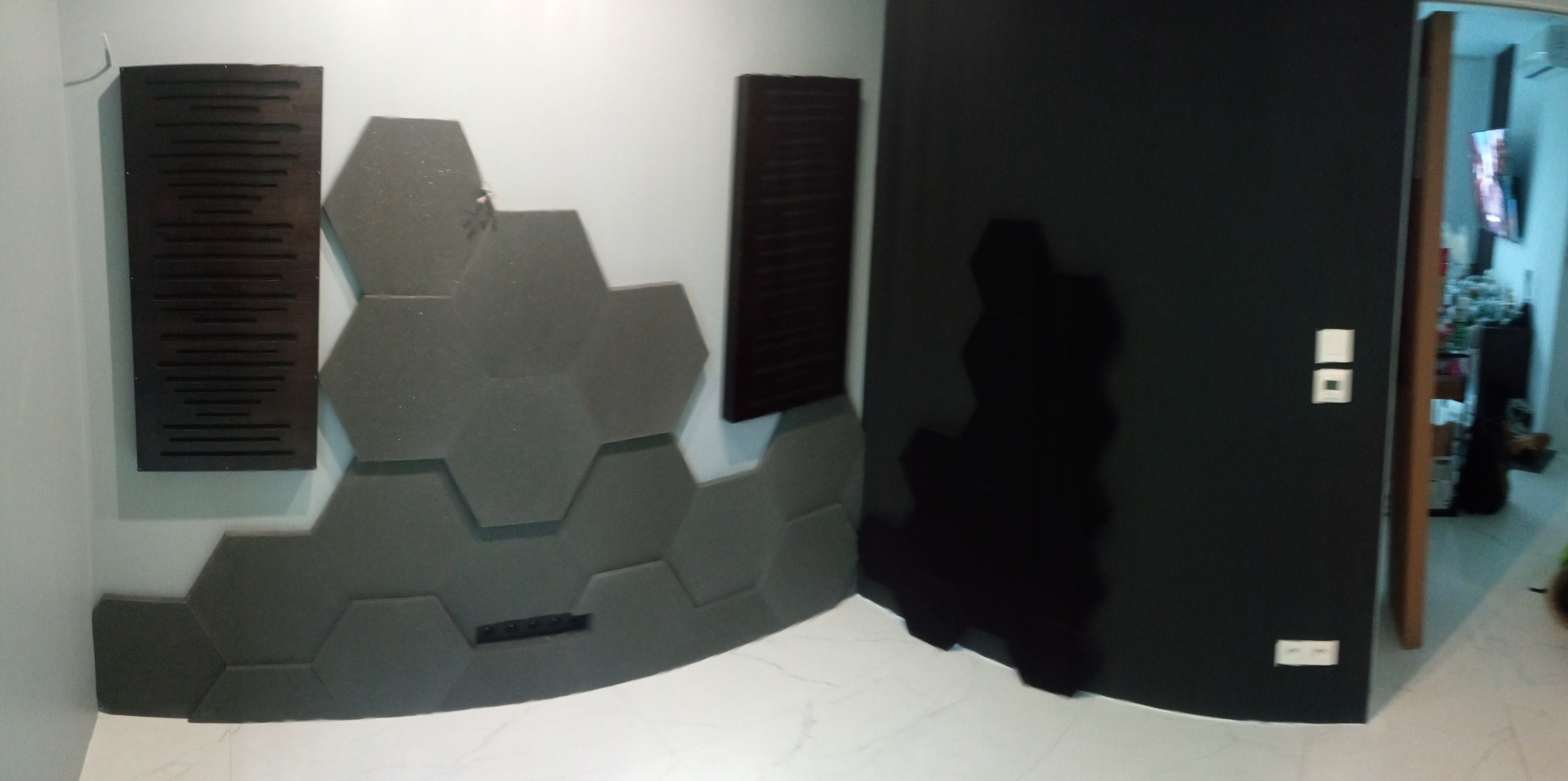 Nowoczesna aranżacja ściany z szarych paneli akustycznych w kształcie heksagonów oraz ciemnych paneli lamelowych, uzupełniona o czarną ścianę z widoczną instalacją elektryczną i fragmentem wnętrza...