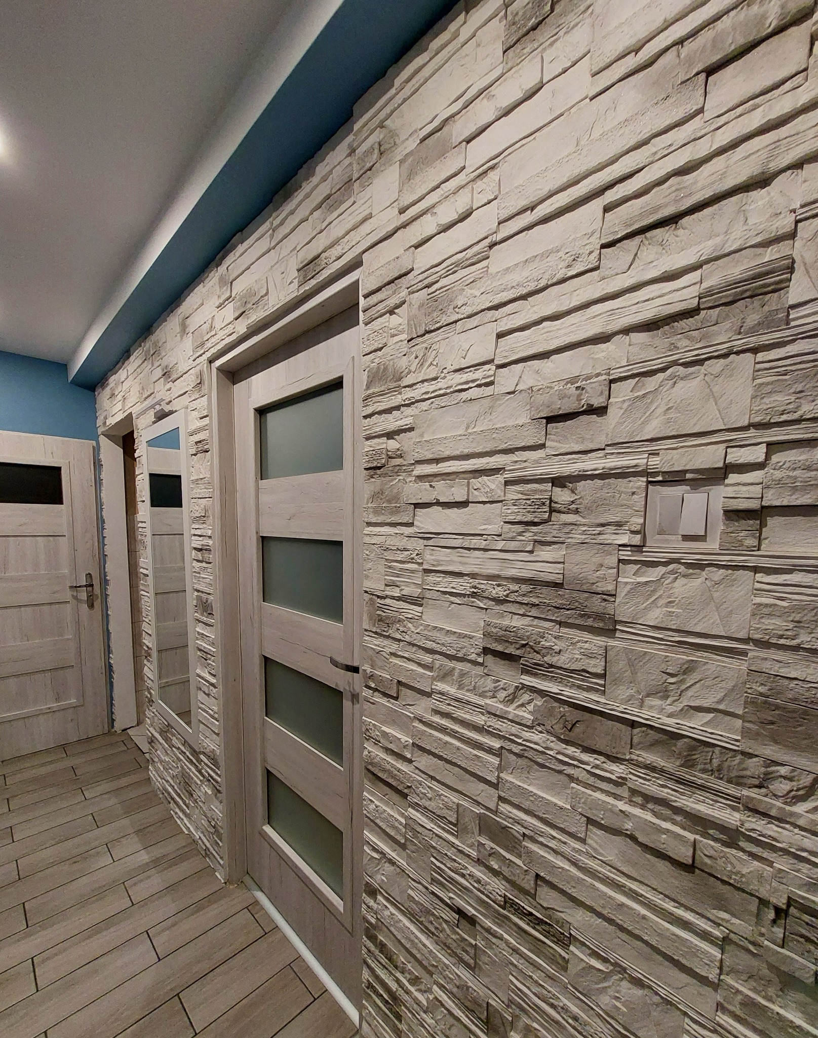 Korytarz z dekoracyjną ścianą z imitacji kamienia w odcieniach szarości, drewnopodobną podłogą z płytek i dwiema drewnianymi drzwiami z przeszkleniami, widok na włącznik światła na kamiennej ścianie.