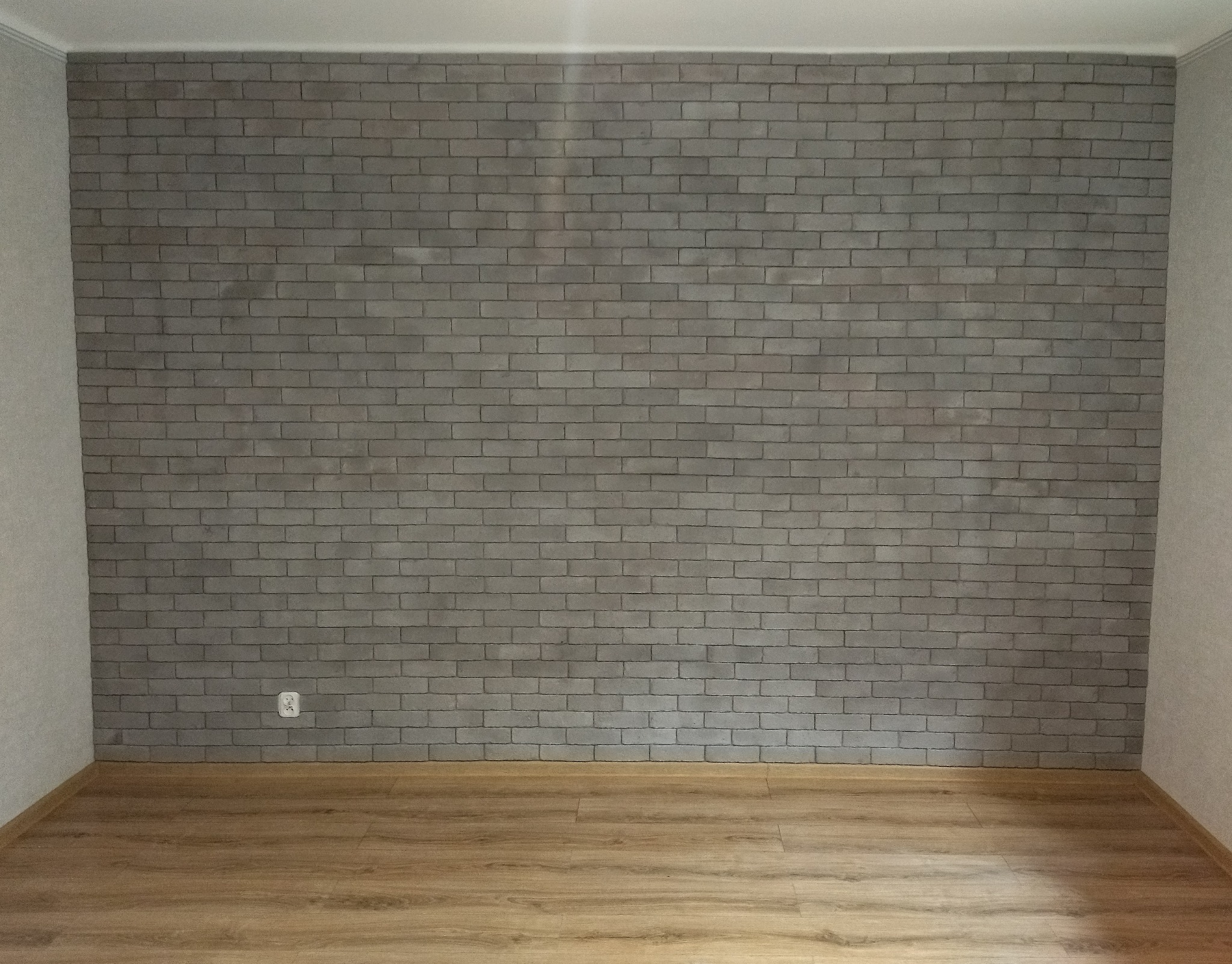 Ściana wykończona szarą cegłą dekoracyjną z widocznym gniazdkiem elektrycznym na dole, podłoga wyłożona panelami w odcieniach brązu, jasne ściany boczne.
