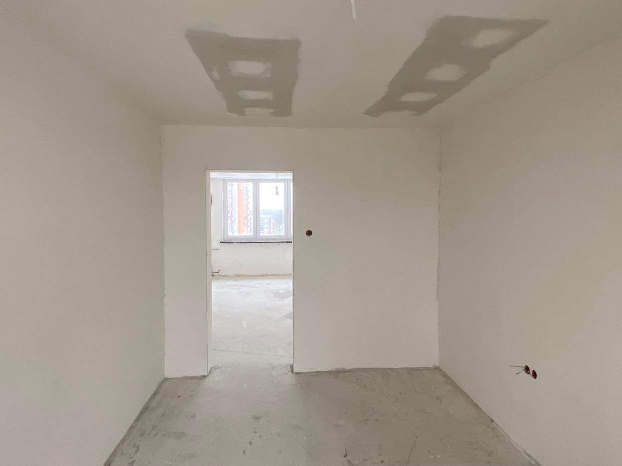Wnętrze pokoju w stanie deweloperskim z widocznymi śladami po szpachlowaniu na suficie i ścianach, surowa podłoga, widok na okno w sąsiednim pomieszczeniu.