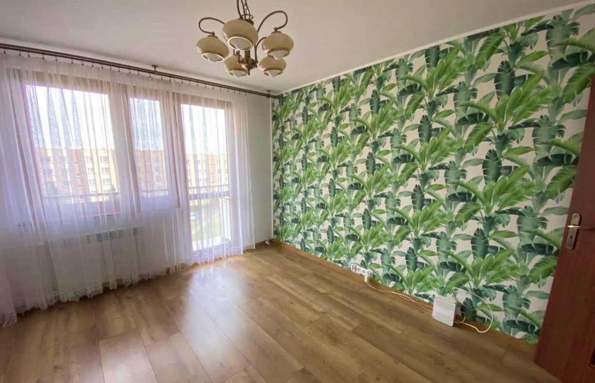 Pokój po odświeżeniu, ściana z wzorem tropikalnych liści, panele podłogowe, okno z białymi firankami, kremowy żyrandol z kilkoma kloszami.