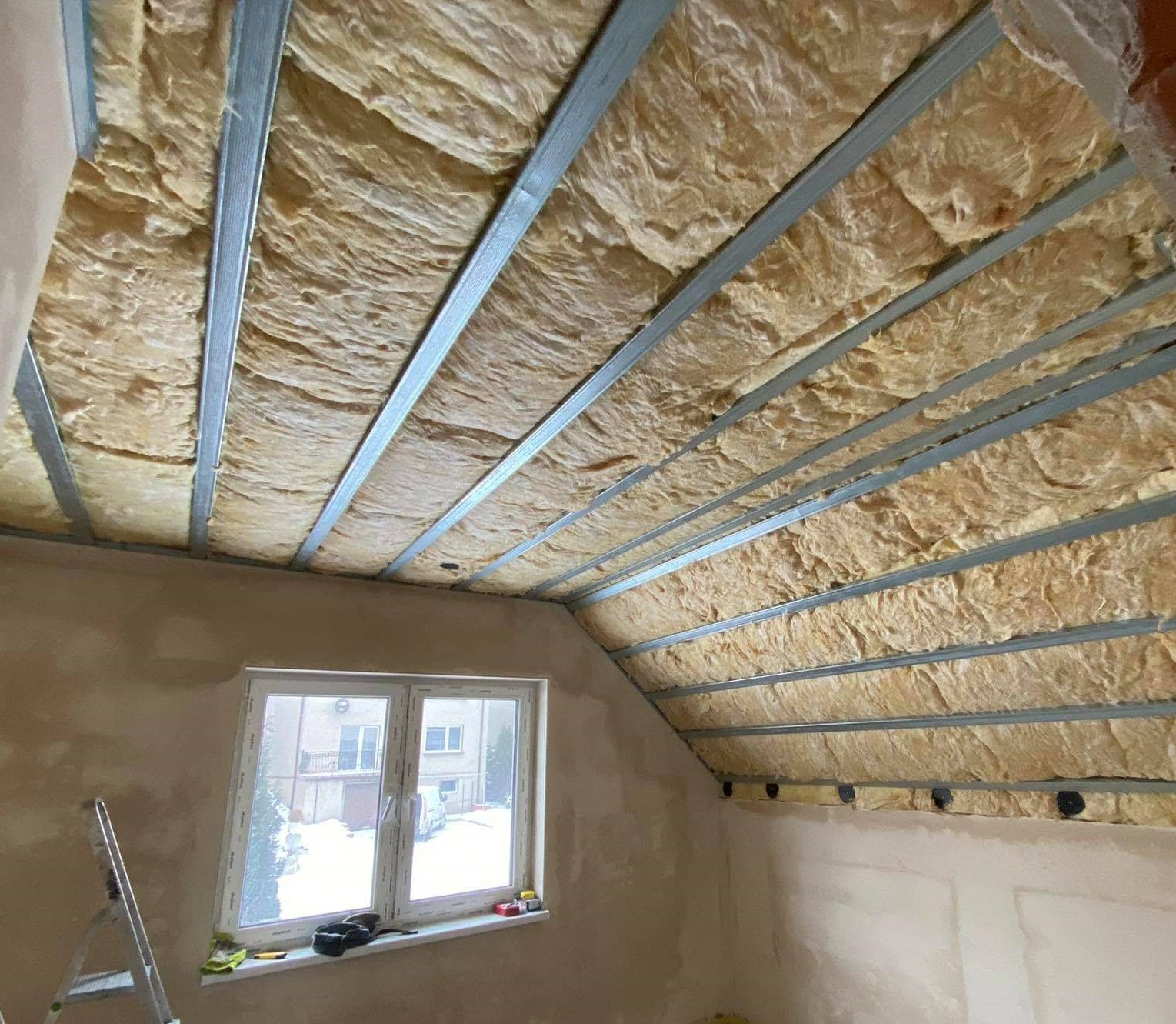 Poddasze w trakcie izolacji termicznej, widoczne metalowe profile konstrukcyjne i wełna mineralna, ściany częściowo otynkowane, w tle okno z widokiem na zaśnieżone otoczenie.