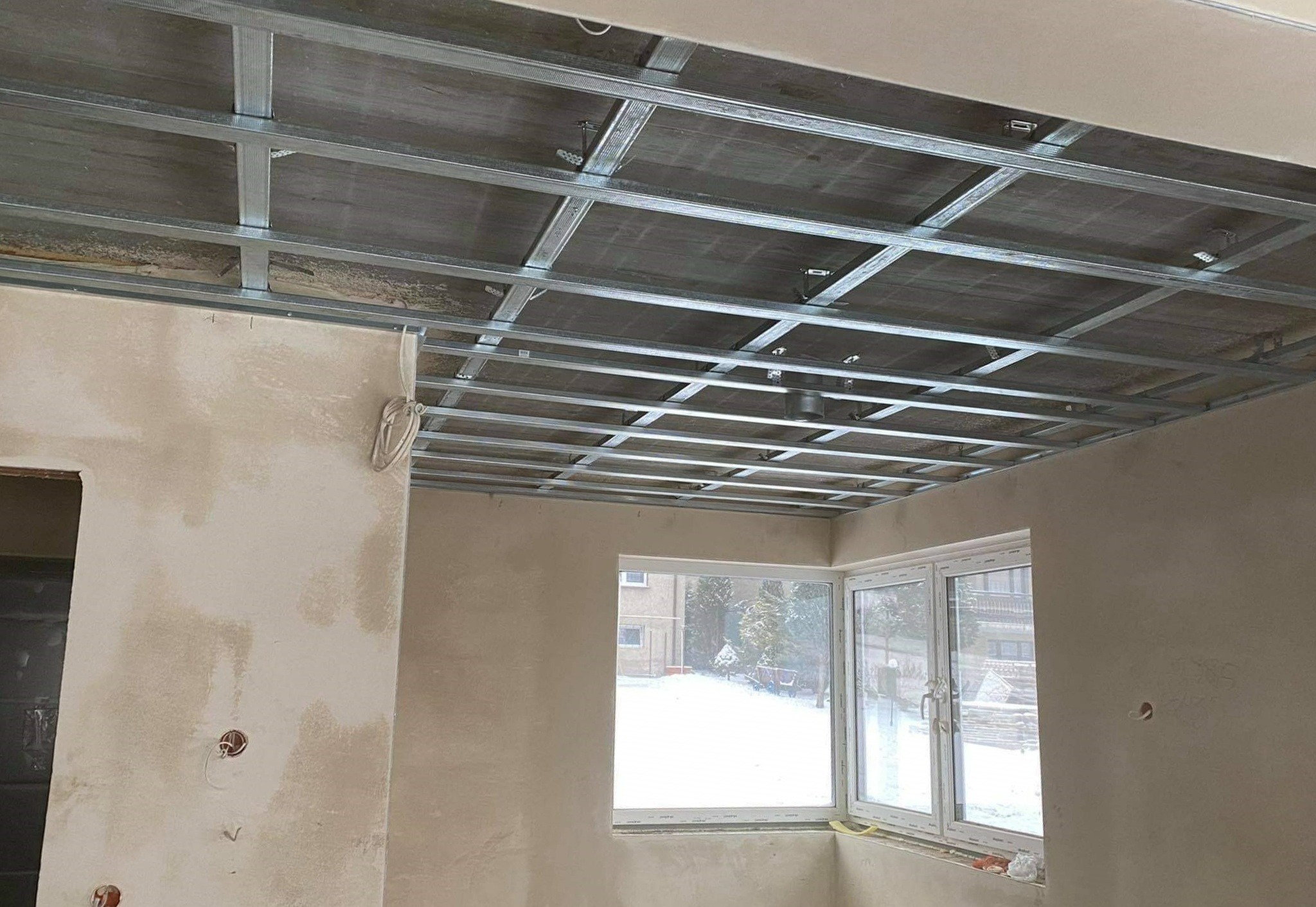 Sufit podwieszany na metalowej konstrukcji w trakcie montażu, ściany w szpachli, widoczne okno z zimowym krajobrazem.