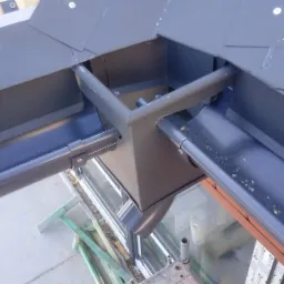 Szczegółowe ujęcie narożnego połączenia grafitowych rynien dachowych z elementami dachu, widoczne zanieczyszczenia wewnątrz rynny, fragment okna i rusztowania w tle.