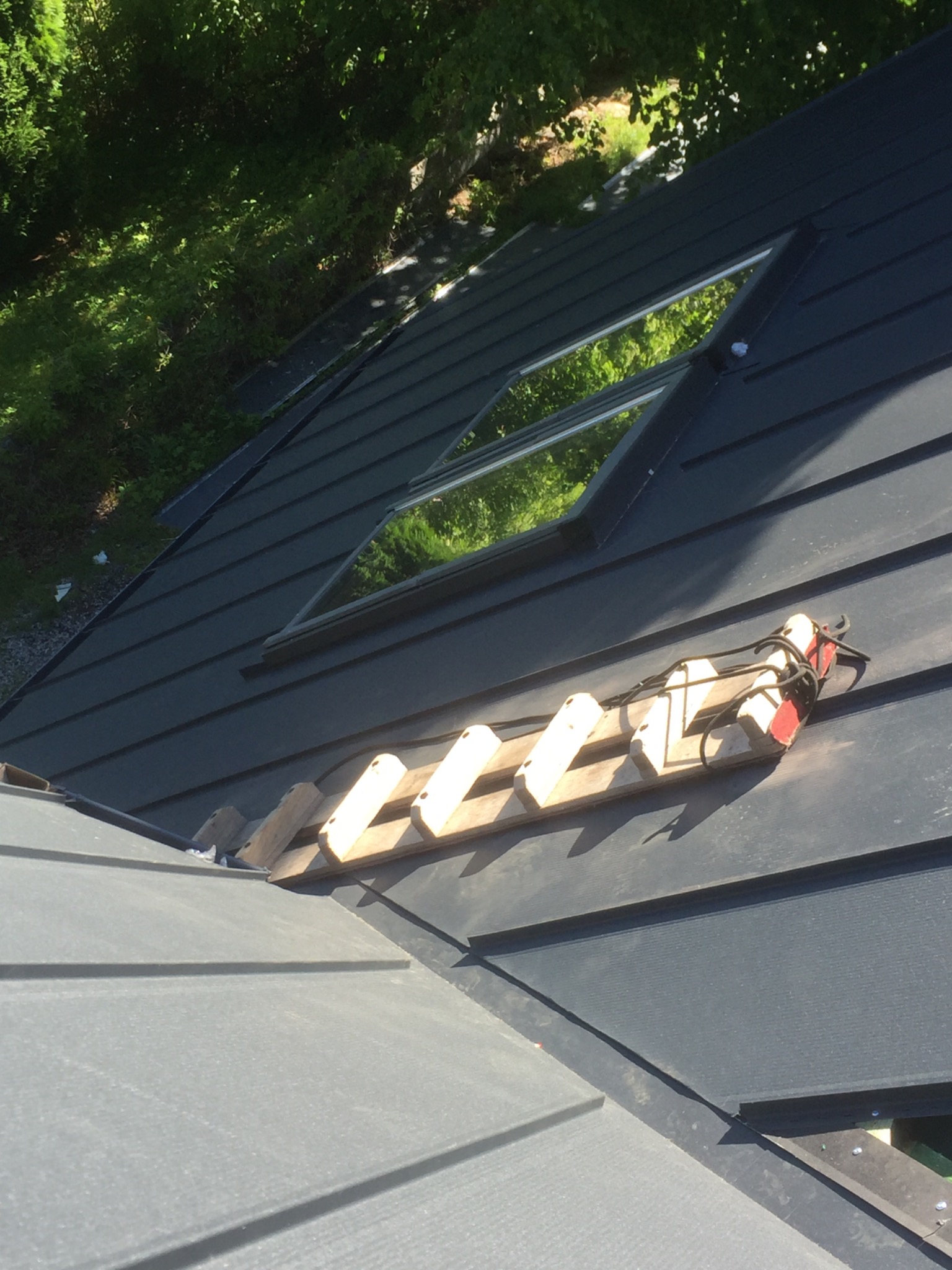 Nowy, grafitowy dach z oknem połaciowym i drabinką dekarską zabezpieczoną linami, widok z góry na fragment dachu w słoneczny dzień.