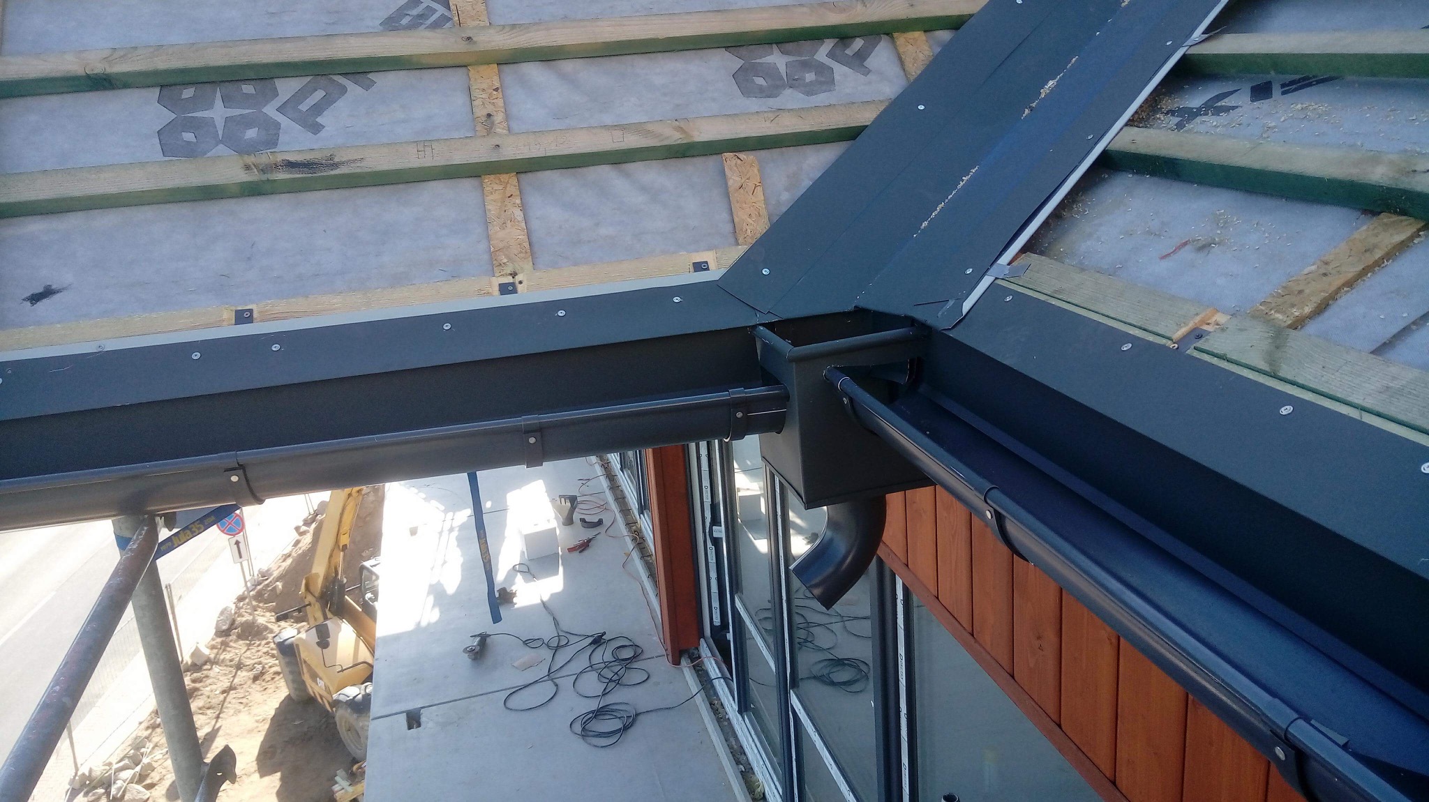 Widok z góry na zamontowane, ciemne orynnowanie i elementy wykończeniowe dachu na tle membrany dachowej i drewnianej konstrukcji więźby; w tle widoczny plac budowy z koparką.