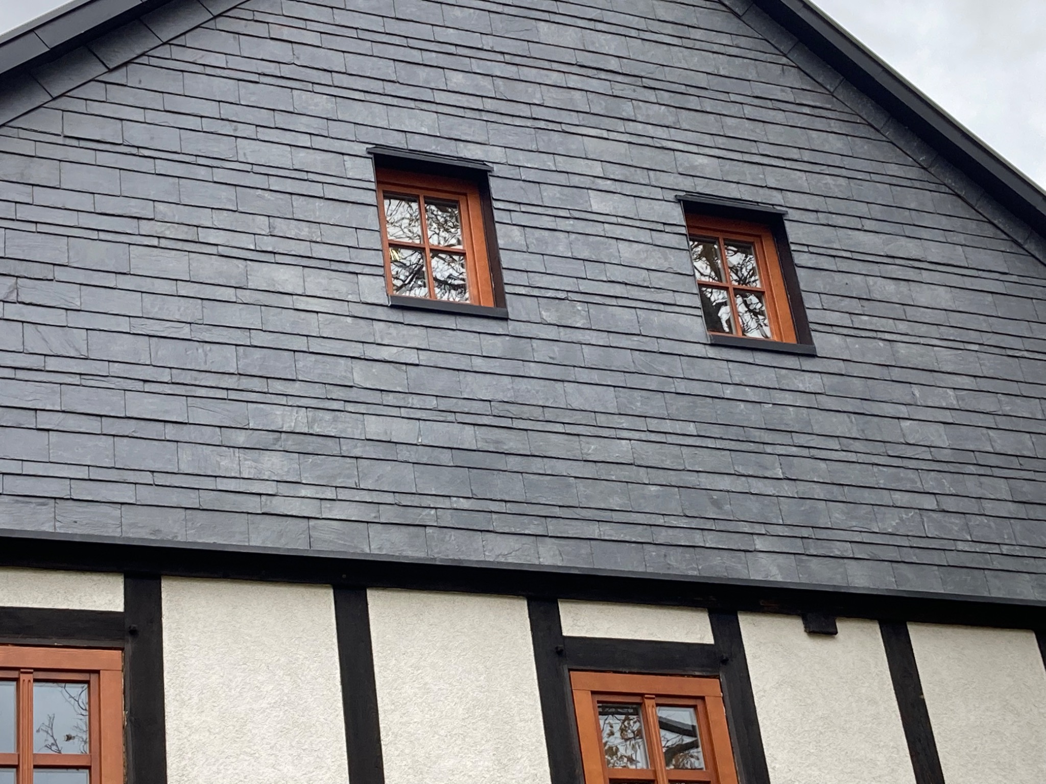 Fragment domu z dachem pokrytym szarym łupkiem, widoczne dwa okna dachowe z brązowymi ramami i fragment ściany szkieletowej z białym wypełnieniem i czarnymi belkami