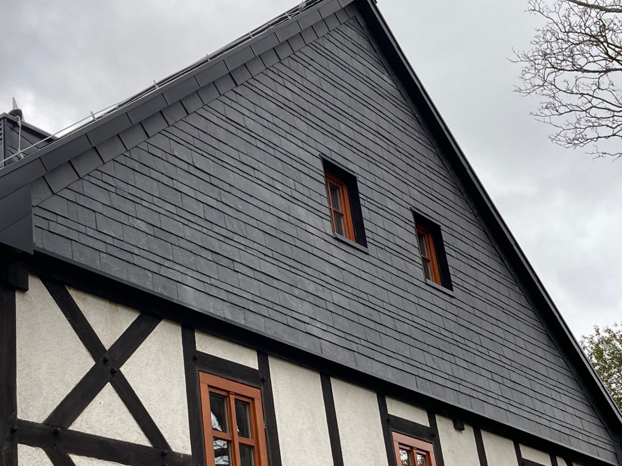 Ciemnoszary dach z łupka na budynku szachulcowym z brązowymi oknami, widok z dołu w pochmurny dzień.