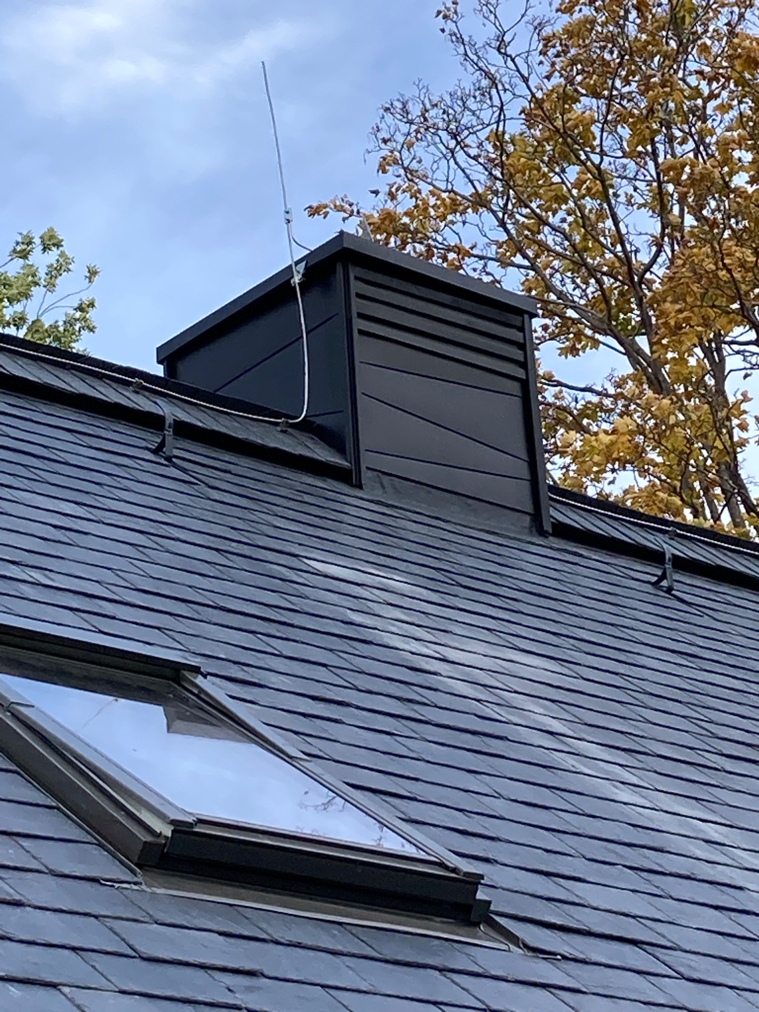 Czarny komin wentylacyjny z anteną na dachu pokrytym ciemnoszarą dachówką łupkową, widoczne okno dachowe i drzewo z żółtymi liśćmi na tle błękitnego nieba.