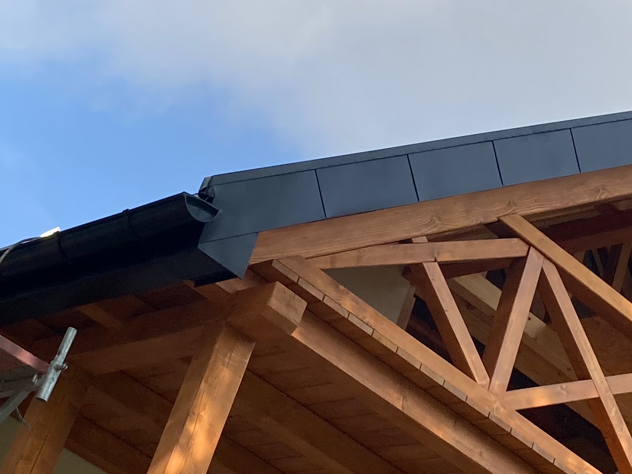 Fragment dachu z czarnymi panelami i rynną, widoczny na tle niebieskiego nieba, z elementami drewnianej konstrukcji więźby dachowej w dolnej części kadru, częściowo zasłoniętej rusztowaniem.