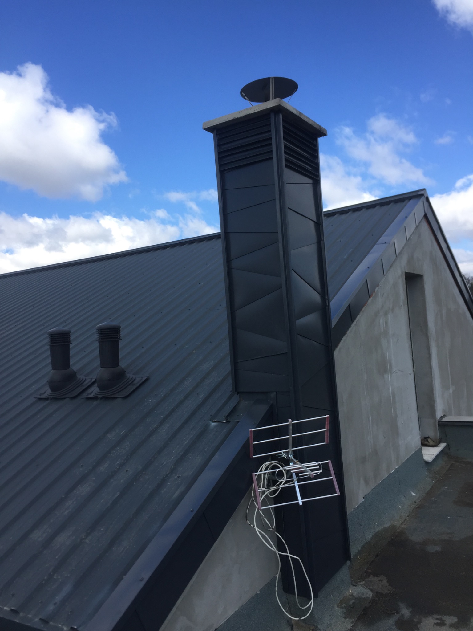 Szary dach z blachy trapezowej z dwoma odpowietrznikami, kominem z metalową obudową i anteną telewizyjną na tle błękitnego nieba z chmurami.