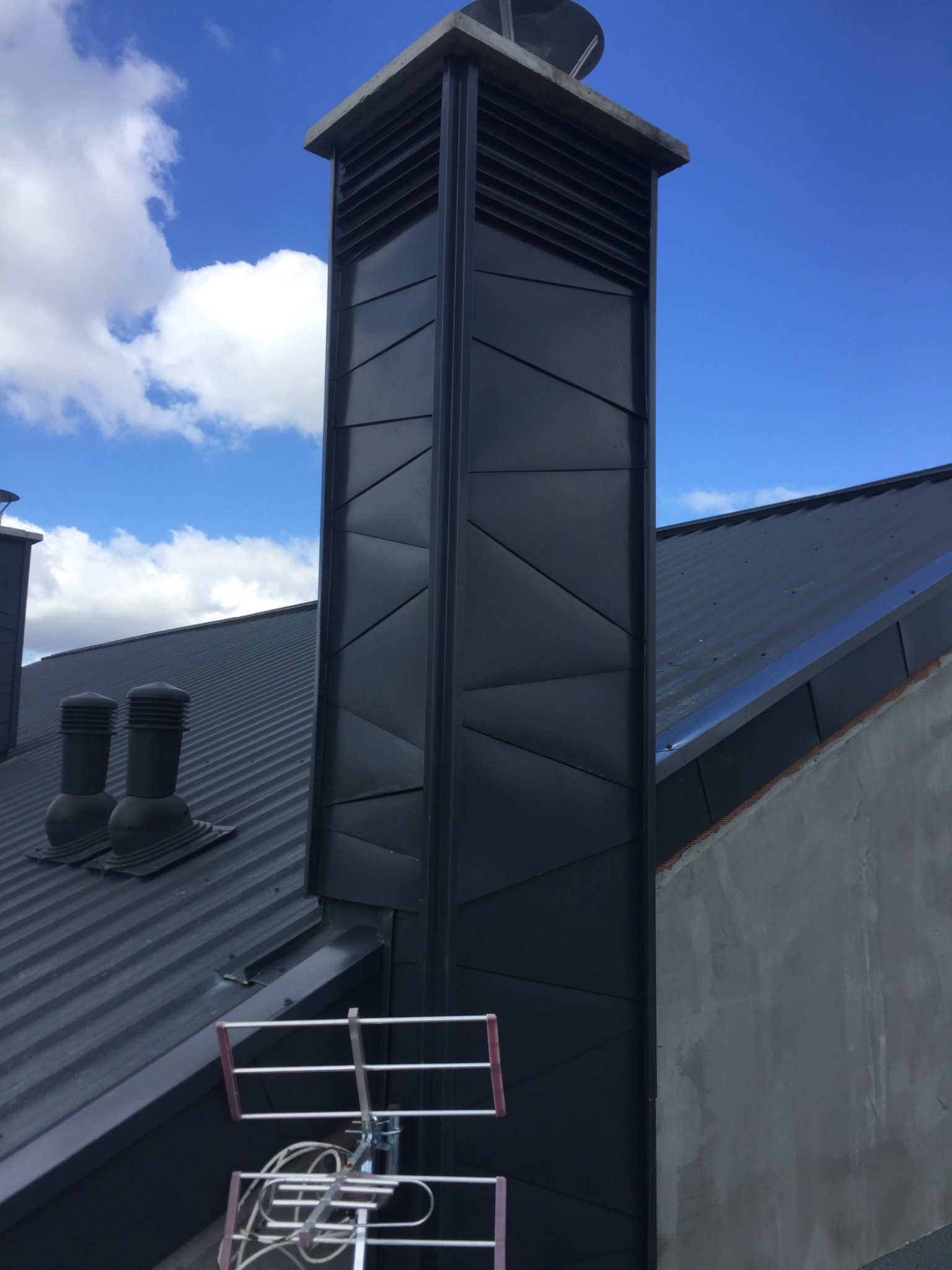 Szary komin z wentylacją na dachu z blachy trapezowej, widoczna antena telewizyjna i fragment ściany budynku.