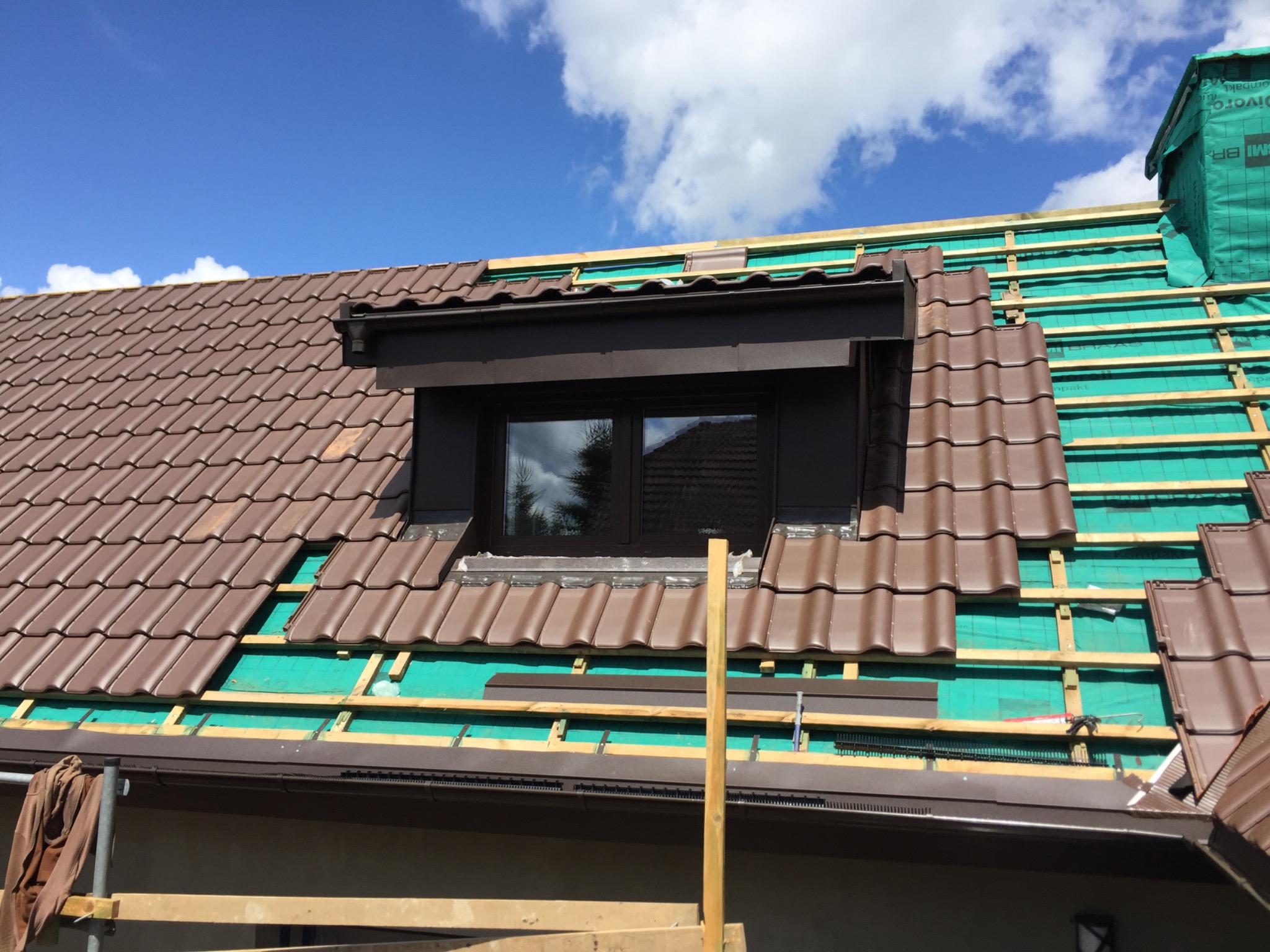Montaż brązowej dachówki wokół okna dachowego, widoczna zielona membrana dachowa i łaty, fragment orynnowania, błękitne niebo z chmurami w tle.
