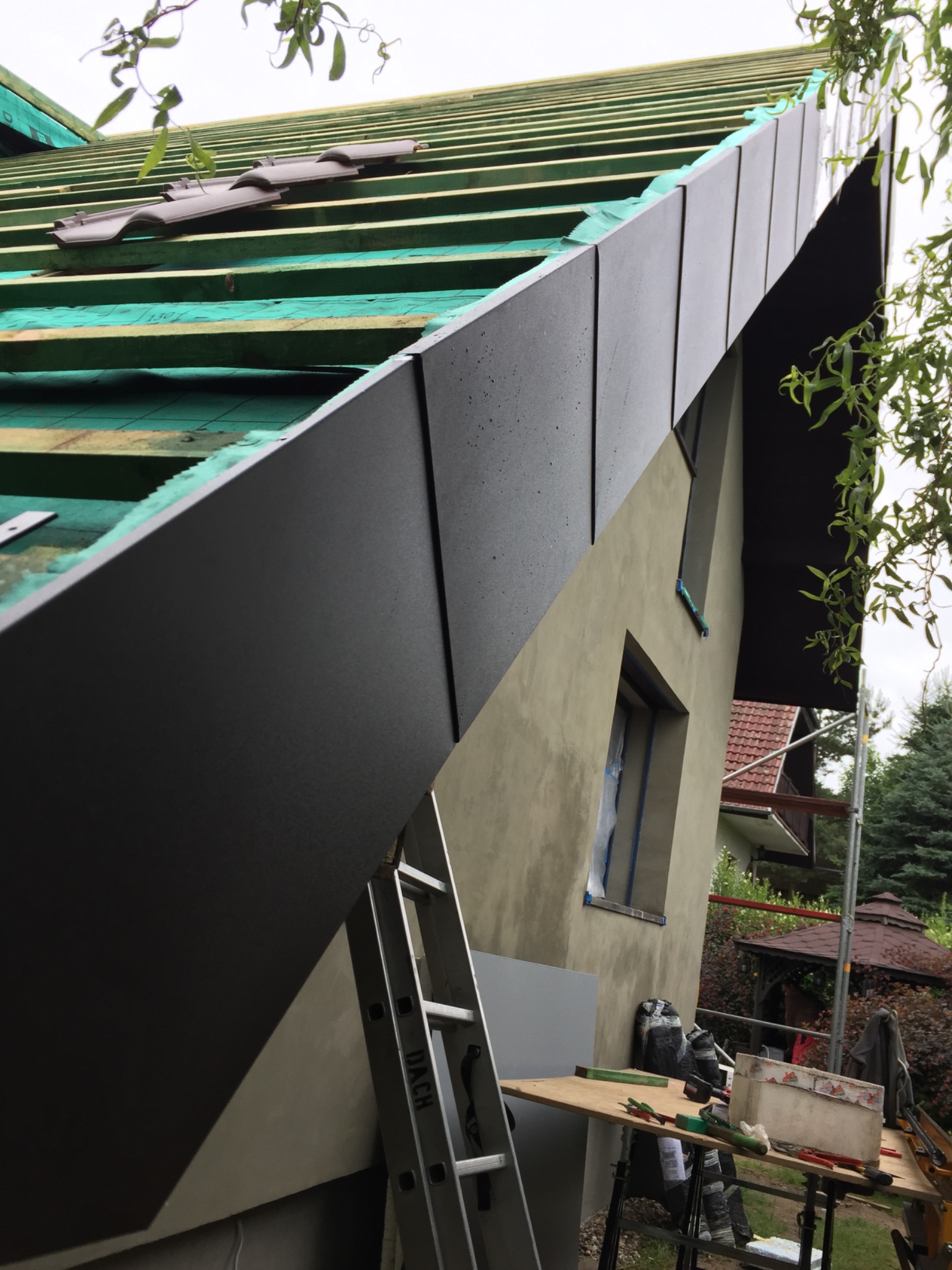 Montaż obróbek blacharskich na dachu domu jednorodzinnego, widoczne deski, folia dachowa oraz częściowo ułożona dachówka, drabina i stół z narzędziami.