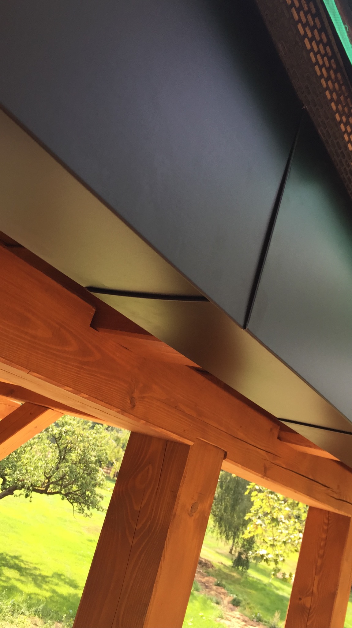 Spojrzenie z dołu na fragment dachu z ciemnej blachy trapezowej, widoczne drewniane elementy konstrukcyjne oraz fragment zielonego ogrodu w tle.