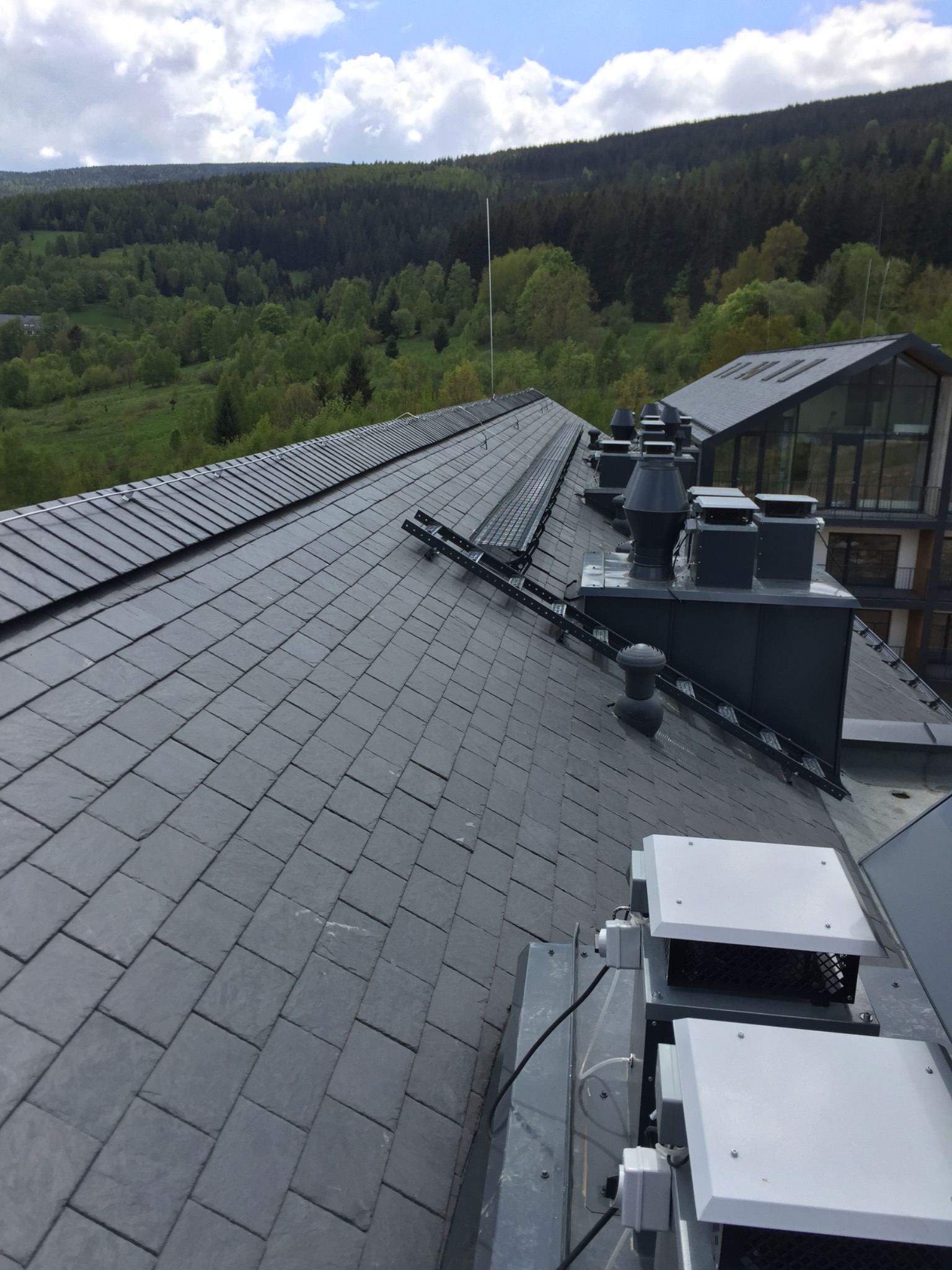 Dach pokryty ciemnoszarą łupkiem z widocznymi kominami wentylacyjnymi i konstrukcją wspornikową, w tle zalesione wzgórza.