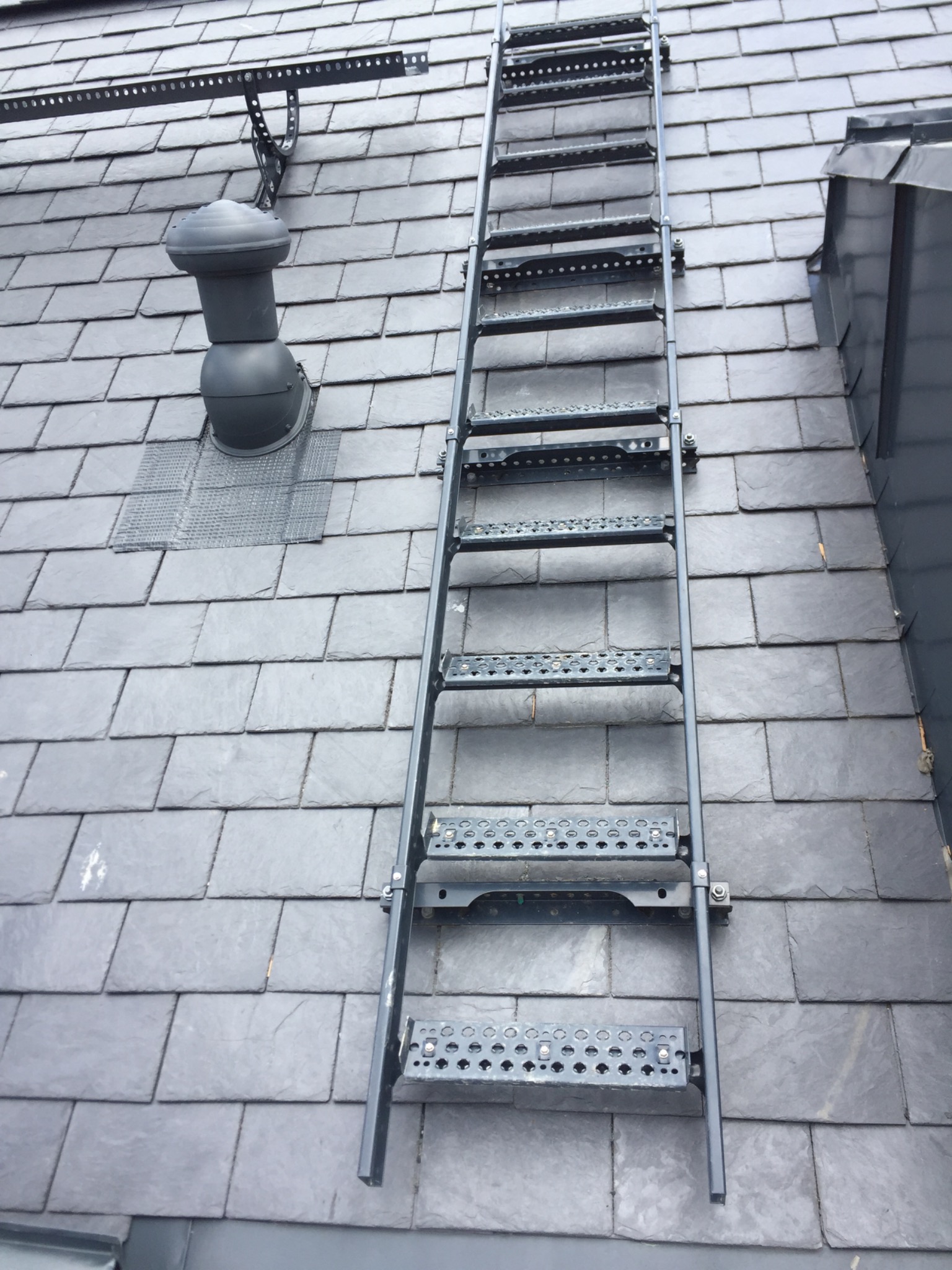 Stroma, grafitowa połać z łupka naturalnego z zamontowaną metalową drabinką dachową i kominkiem wentylacyjnym z osłoną.