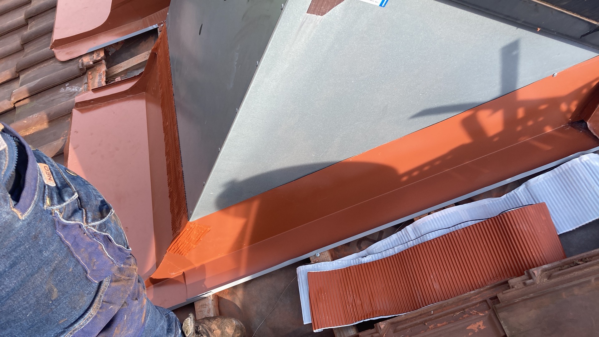 Montaż obróbek blacharskich na dachu z wykorzystaniem blachy w kolorze ceglastym i szarym, widoczne elementy mocujące oraz fragment ubrania roboczego montera.