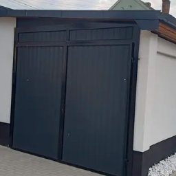 Patryk Żurek usługi spawalnicze MT-spaw - Nowoczesny garaż z dwuskrzydłową, antracytową bramą segmentową, białą elewacją i czarną podmurówką, widok z zewnątrz na tle posesji.
