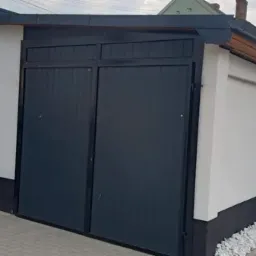 Nowoczesny garaż z dwuskrzydłową, antracytową bramą segmentową, białą elewacją i czarną podmurówką, widok z zewnątrz na tle posesji.