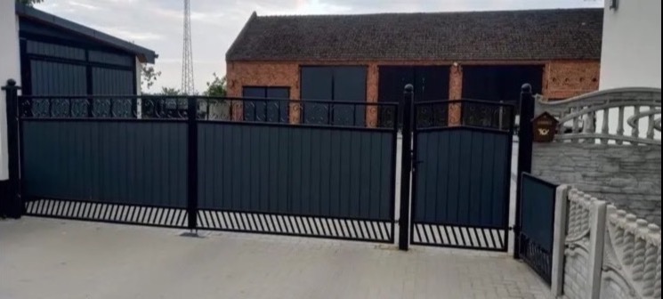 Nowoczesna, grafitowa brama wjazdowa z dekoracyjnymi elementami, widok na posesję z ceglanym budynkiem w tle.