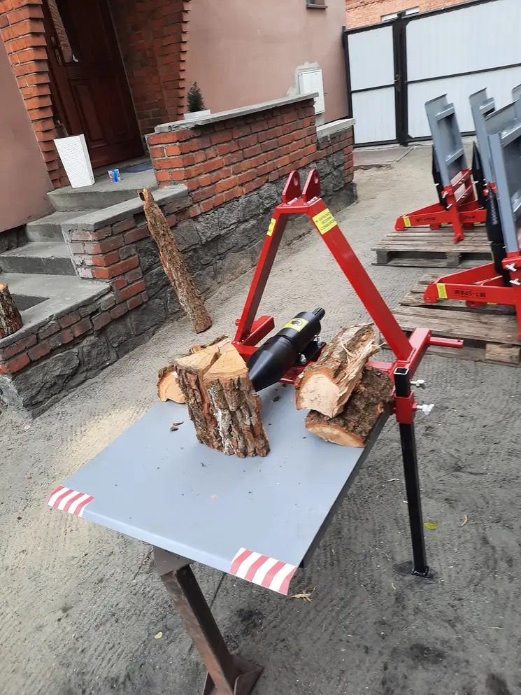 Hydrauliczna łuparka do drewna z czerwonymi elementami konstrukcyjnymi i czarnym cylindrem, ustawiona na szarym stole z paskami ostrzegawczymi, z przygotowanym drewnem do łupania, w tle budynek...