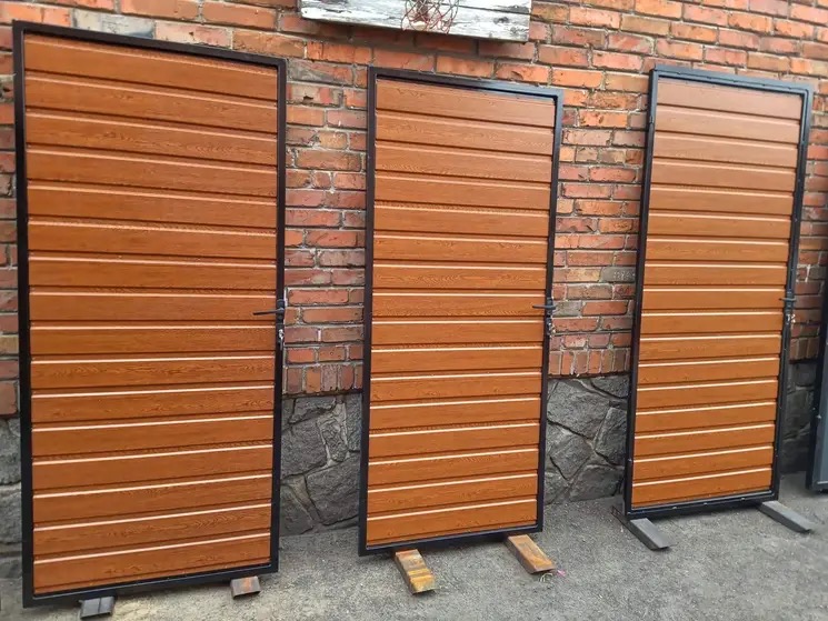 Trzy brązowe bramy z poziomymi panelami imitującymi drewno, z czarną ramą, ustawione na drewnianych klockach przed ceglaną ścianą.