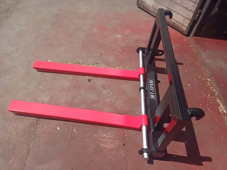 Widok z przodu na nowy, czarny wózek widłowy z czerwonymi widłami, stojący na czerwonej podłodze w pobliżu drewnianej ściany.