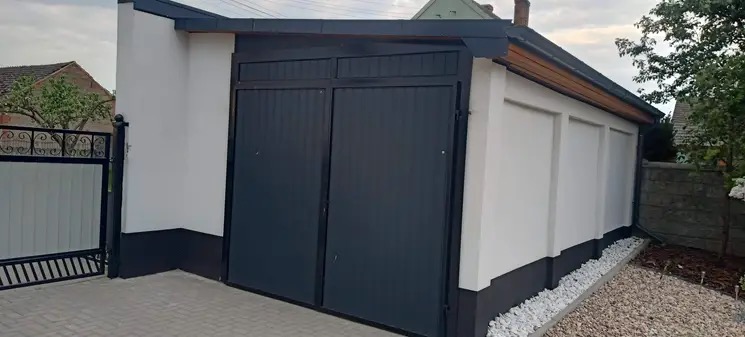 Nowoczesny garaż z dwuskrzydłową, antracytową bramą segmentową, białą elewacją i czarną podmurówką, widok z zewnątrz na tle posesji.