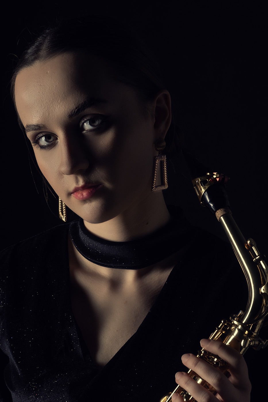 Portret młodej kobiety z saksofonem w ręku, w ciemnym oświetleniu studyjnym. Uwagę przykuwają geometryczne kolczyki i choker.