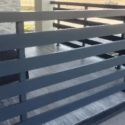 Nowoczesne, poziome, metalowe zabezpieczenie tarasu w kolorze grafitowym, z widocznymi elementami mocującymi, dekoracją w postaci suszonych kwiatów i czerwonej wstążki, oraz fragmentem mebli...