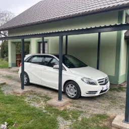 Biały samochód osobowy marki Mercedes-Benz zaparkowany pod nowoczesną wiatą garażową z metalową konstrukcją, widok z zewnątrz na tle domu z zieloną elewacją i brązowym dachem.