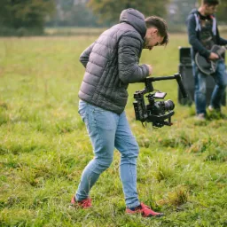 Młody mężczyzna w kurtce i jeansach, pochylony, obsługuje profesjonalną kamerę z rigiem na łące, w tle muzyk z gitarą elektryczną i wzmacniaczem.