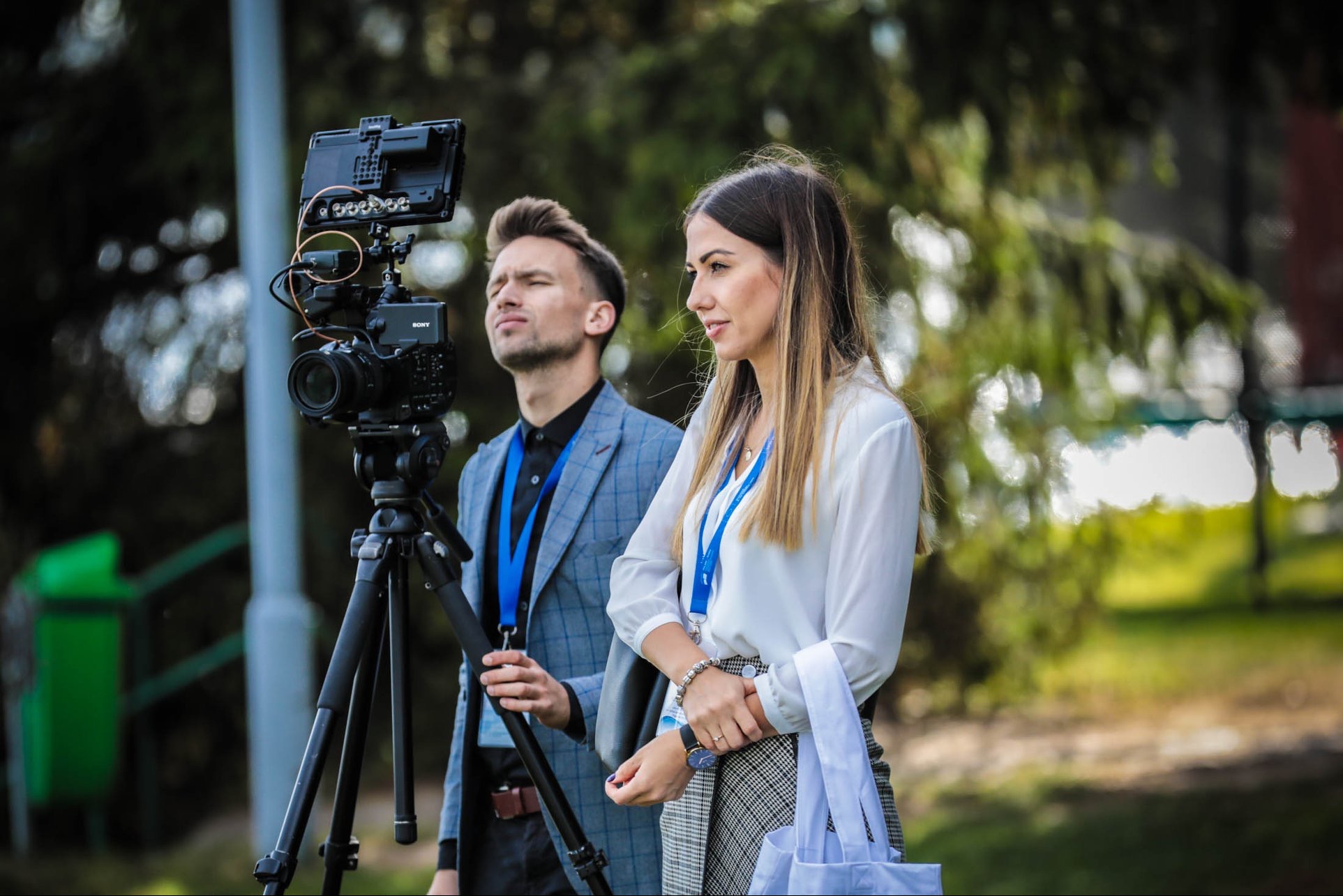 Kamerzysta z asystentką podczas kręcenia sceny plenerowej, widoczny profesjonalny sprzęt filmowy na statywie oraz identyfikatory na szyjach członków ekipy.
