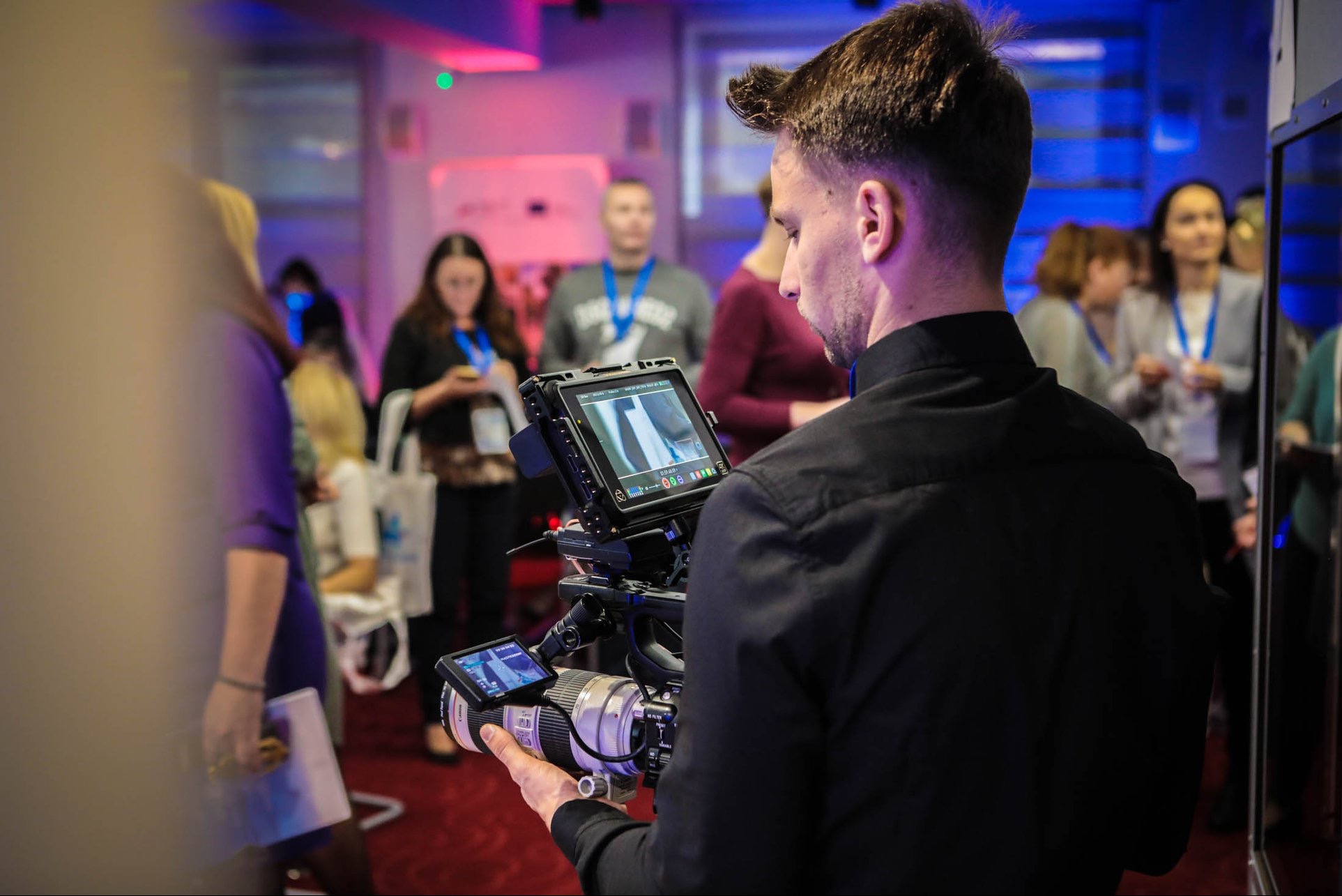 Operator kamery z profesjonalnym sprzętem, w tle uczestnicy wydarzenia podczas filmowania.