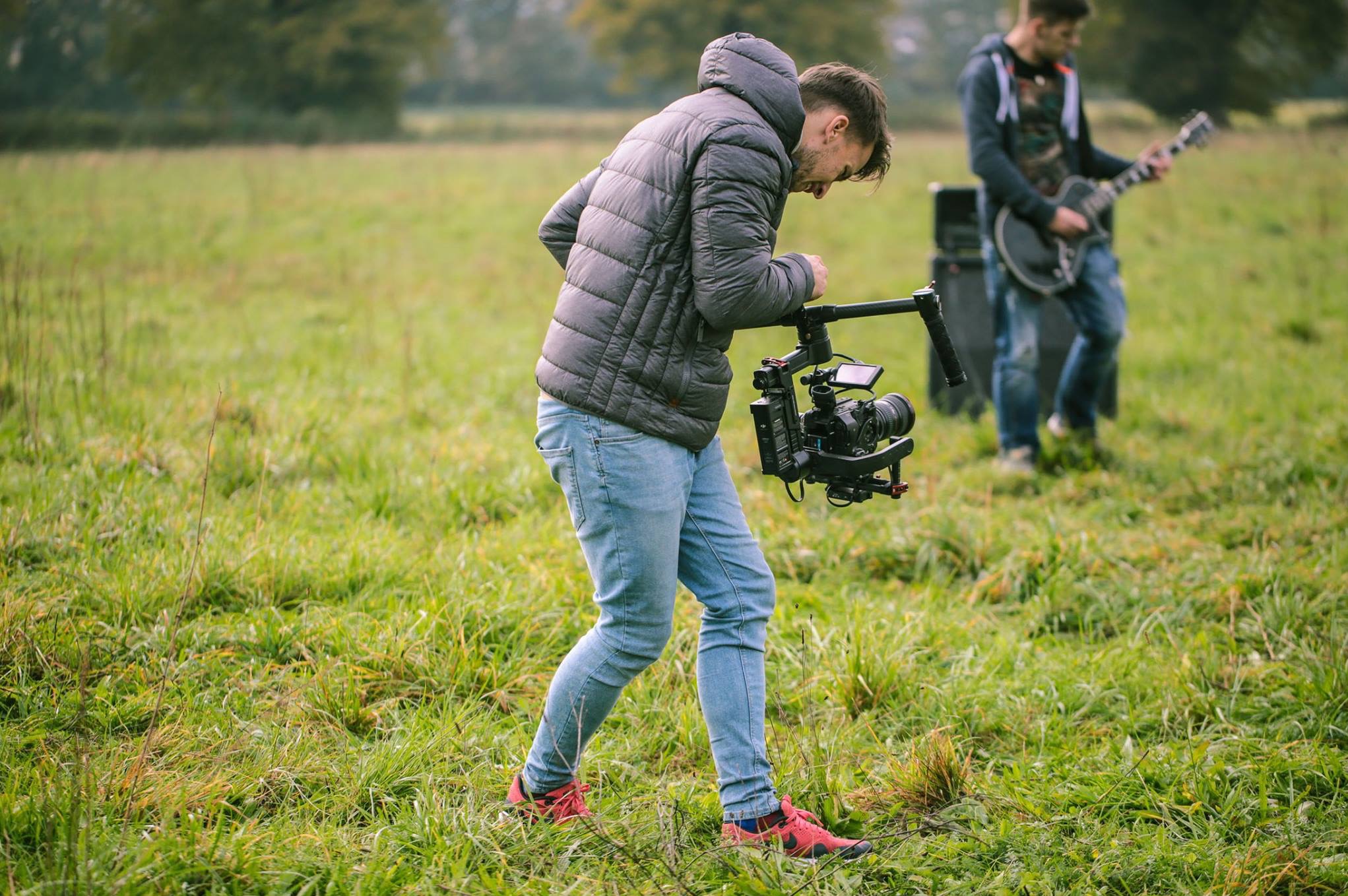 Młody mężczyzna w kurtce i jeansach, pochylony, obsługuje profesjonalną kamerę z rigiem na łące, w tle muzyk z gitarą elektryczną i wzmacniaczem.