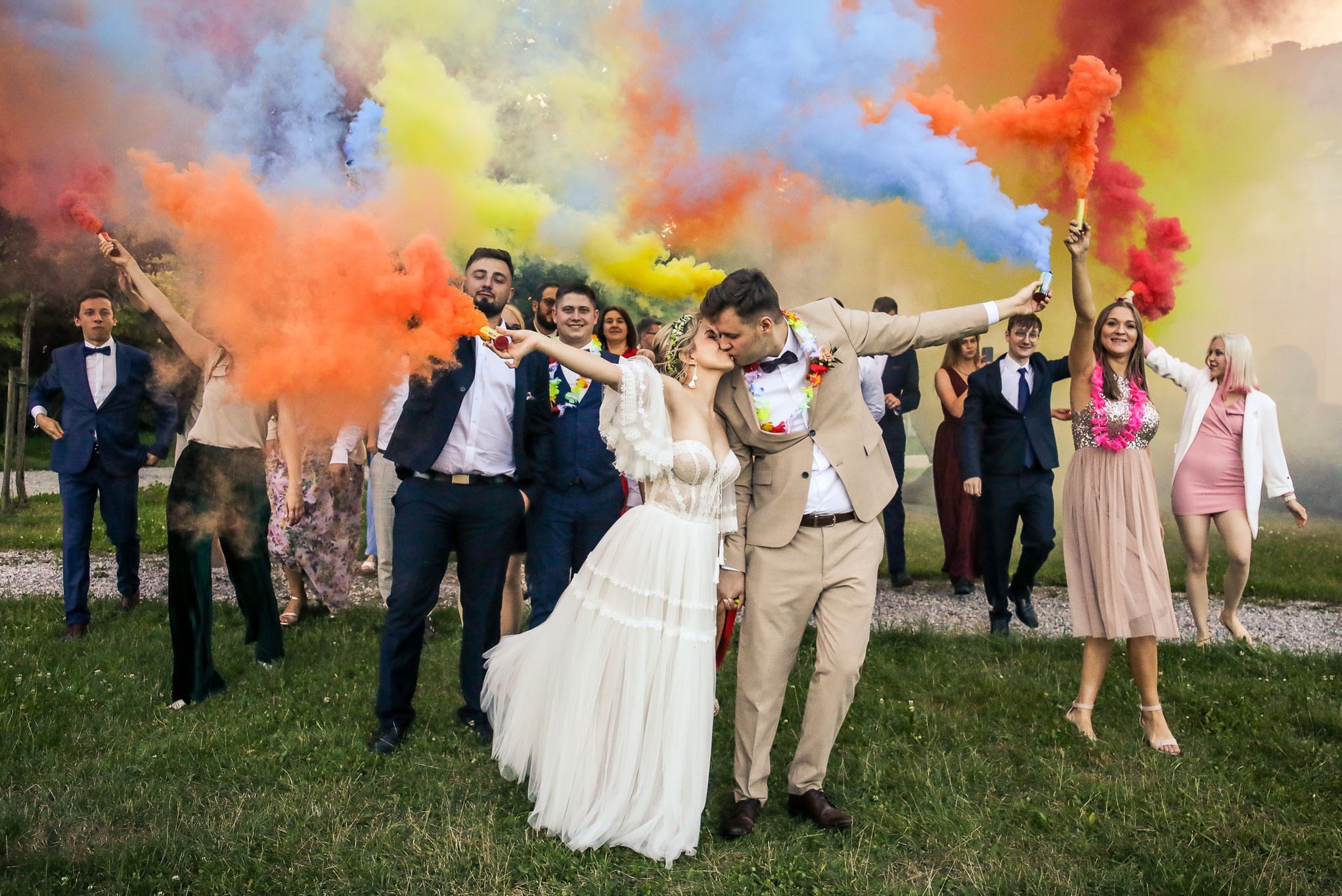 Pocałunek młodej pary na tle kolorowych dymów podczas wesela plenerowego, goście w tle trzymają race dymne.