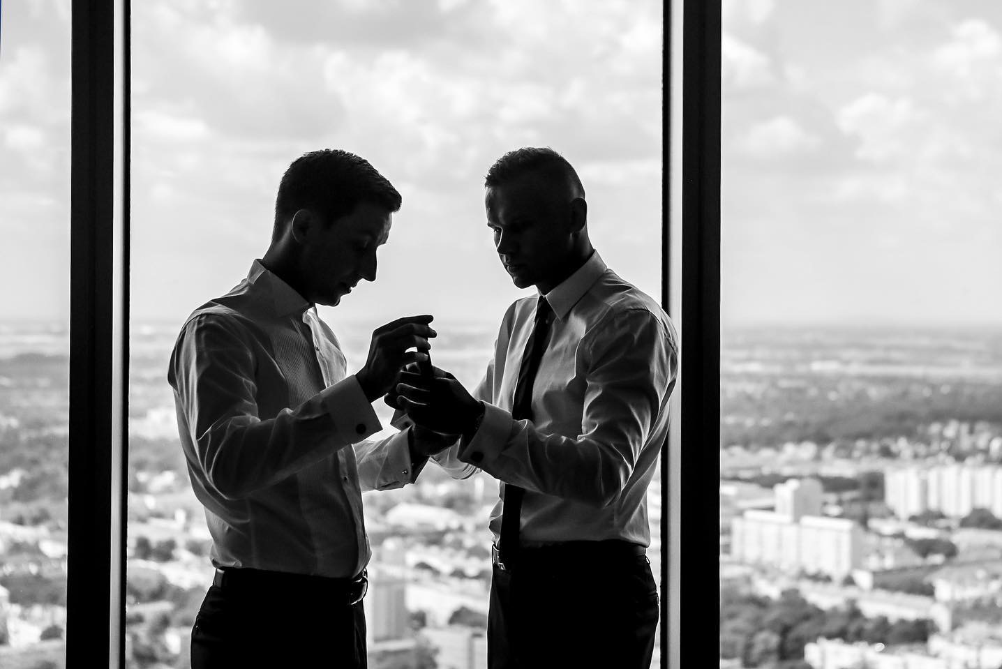 Czarno-białe zdjęcie dwóch mężczyzn w koszulach, stojących przy oknie z widokiem na miasto, prawdopodobnie poprawiających mankiety lub zapinających spinki.