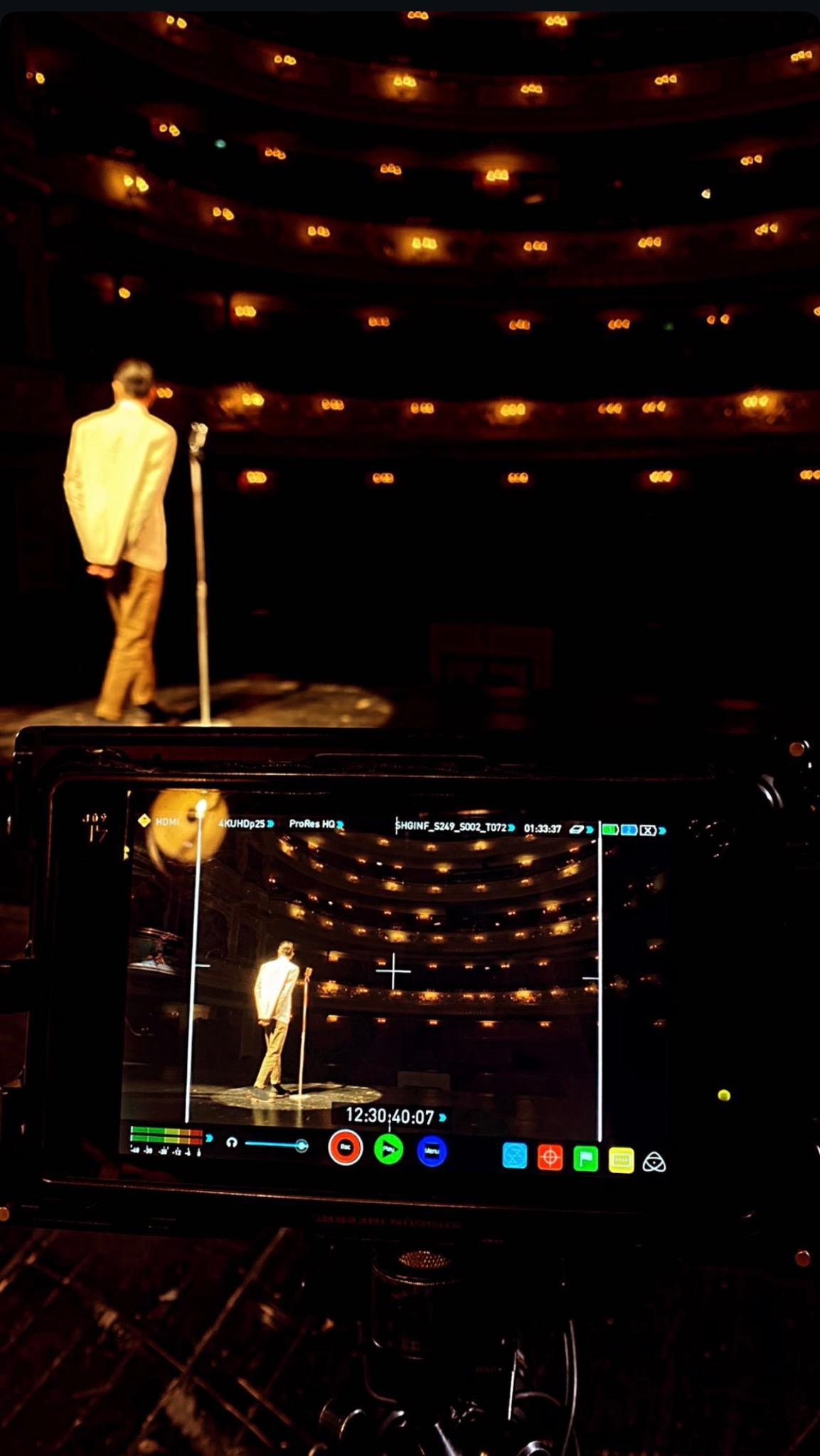 Ekran kamery filmowej rejestruje ujęcie zza kulis: mężczyzna w jasnym garniturze stoi na scenie oświetlonej reflektorem, przed mikrofonem, na tle widowni w teatrze z rzędami świateł.