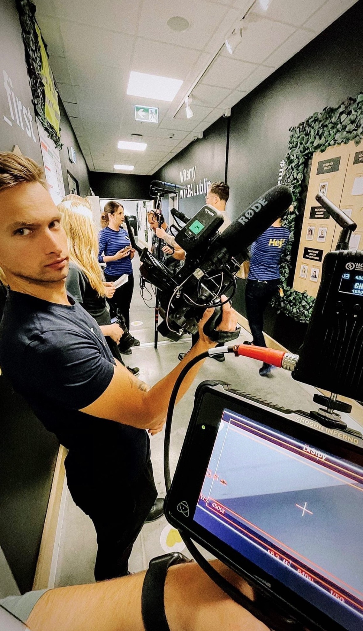 Ekipa filmowa w trakcie nagrywania w korytarzu, widoczny operator kamery z profesjonalnym sprzętem, mikrofon Rode, monitor podglądowy Shogun Inferno oraz osoby w tle.