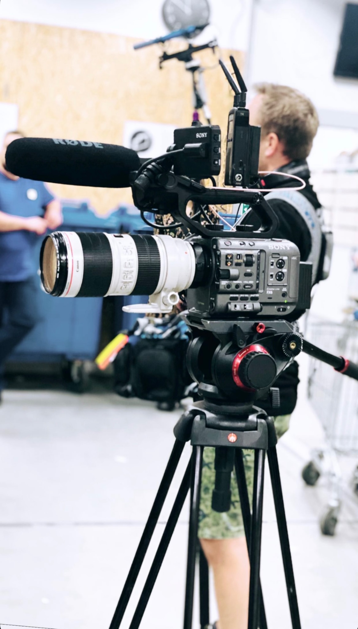 Profesjonalna kamera Sony FX6 z obiektywem Canon i mikrofonem Rode na statywie Manfrotto, w tle osoba i wózki sklepowe.