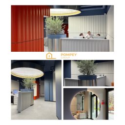 POMPEY GROUP SPÓŁKA Z OGRANICZONĄ ODPOWIEDZIALNOŚCIĄ - Nowoczesne wnętrze biurowe z recepcją, rośliną w donicy i oświetleniem LED. Kolorystyka: biel, granat i pomarańcz. Widoczne osoby za ladą.