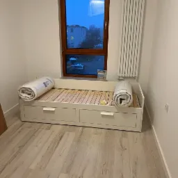 Wnętrze pokoju po remoncie: biała, drewniana rama łóżka z szufladami, drewniany stelaż, dwa zwinięte materace w folii, parkiet, okno z widokiem na budynek, biały grzejnik.