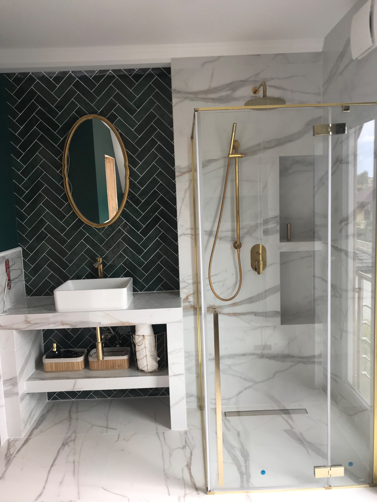 Nowoczesna łazienka z kabiną prysznicową ze złotymi elementami, umywalką nablatową i ścianą wyłożoną ciemnozielonymi płytkami w jodełkę.