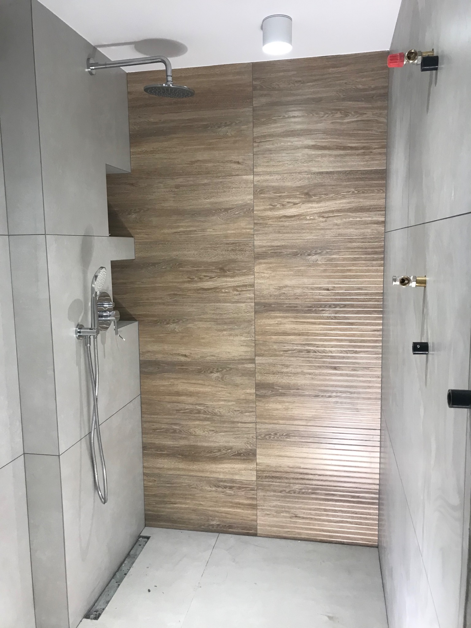Wykończona kabina prysznicowa z szarymi płytkami i drewnopodobną ścianą, widoczny odpływ liniowy w podłodze i podwieszana deszczownica.