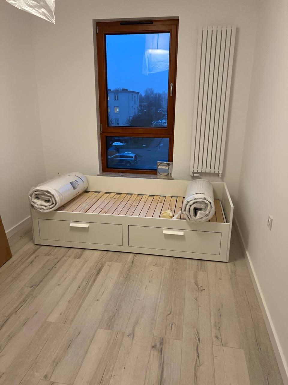 Wnętrze pokoju po remoncie: biała, drewniana rama łóżka z szufladami, drewniany stelaż, dwa zwinięte materace w folii, parkiet, okno z widokiem na budynek, biały grzejnik.
